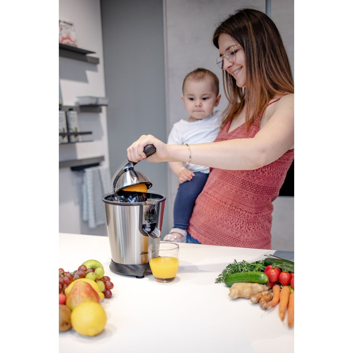 Donna che tiene un bambino, che spremi arance. Vari frutti e verdure sul tavolo.