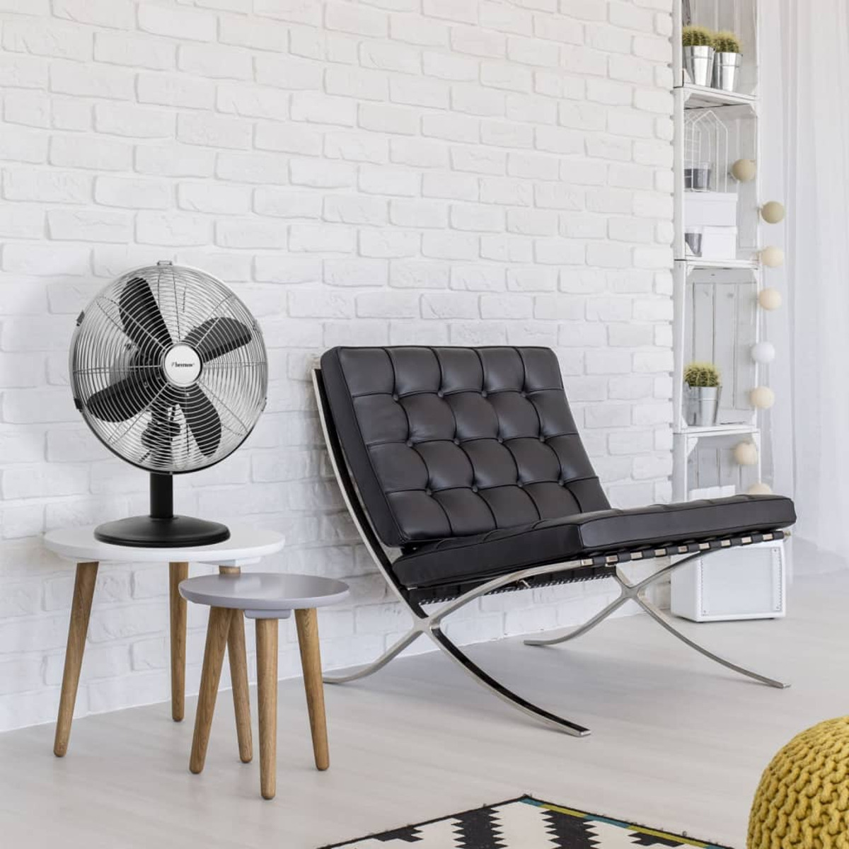 Ventilatore su tavolini, sedia nera, muro di mattoni bianchi, piante e tappeto.