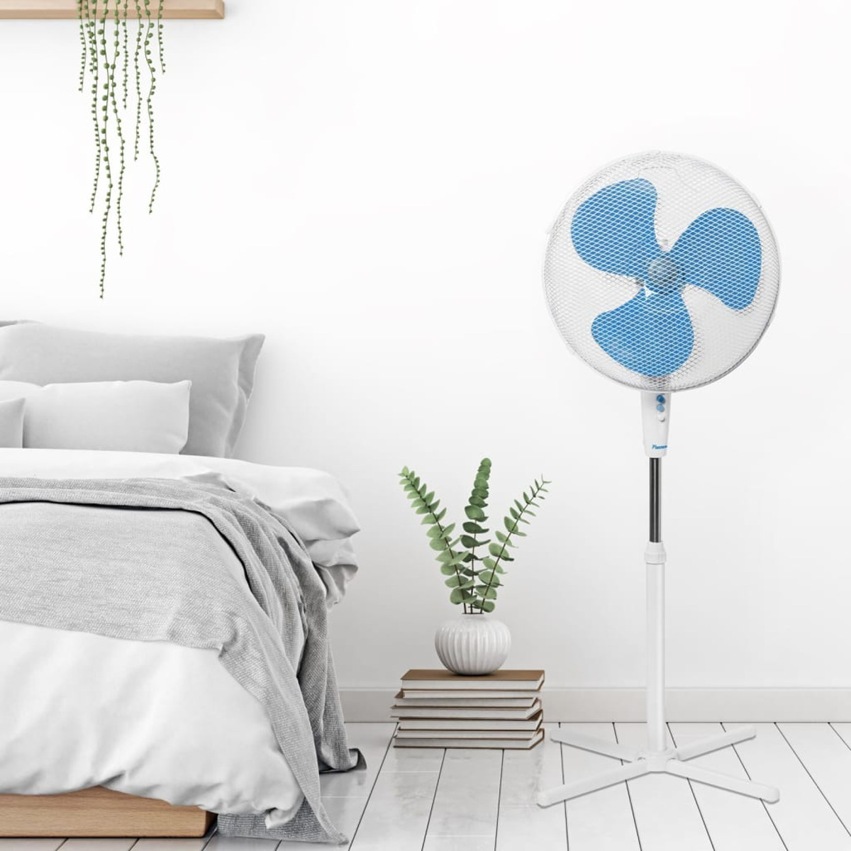 Biały wentylator stojący z niebieskimi łopatkami, obok łóżka i rośliny.