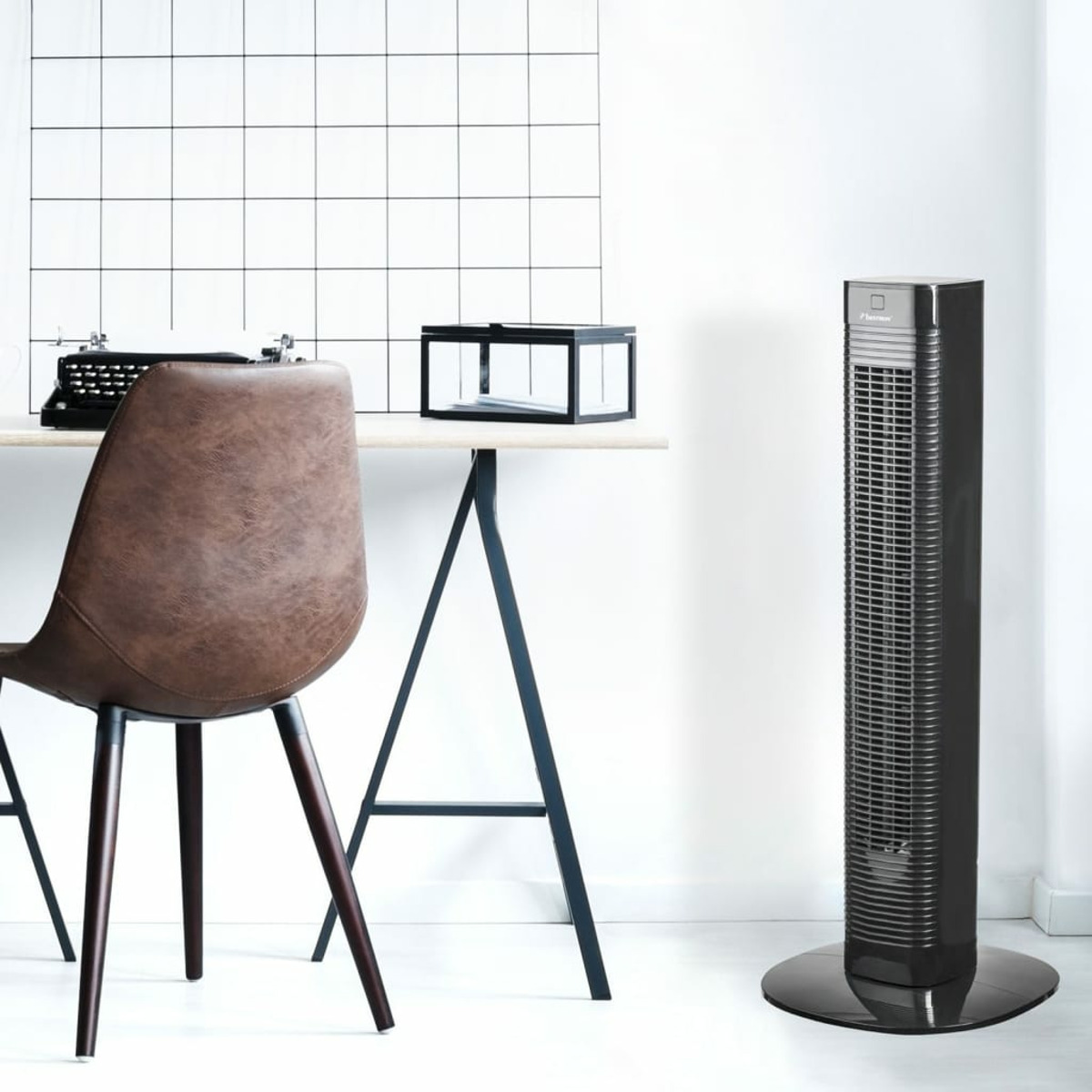 Un ventilatore a torre nero in piedi vicino a una sedia e una scrivania sul muro.
