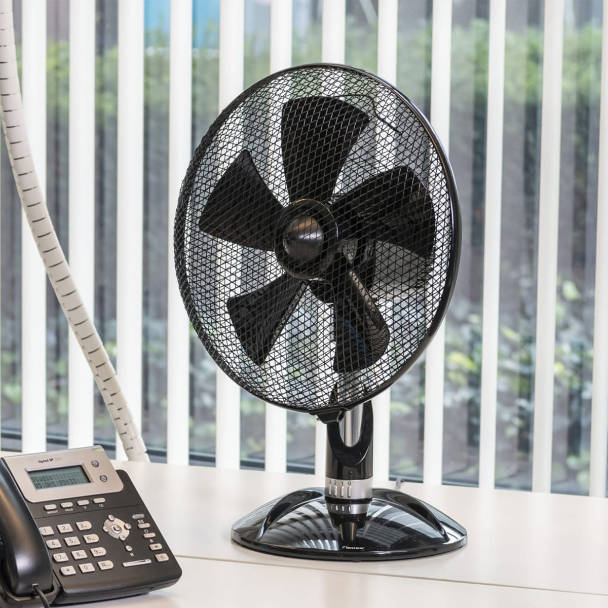 Ventilatore da scrivania nero su scrivania bianca. Dietro, finestra con persiane verticali. Un telefono sulla scrivania.