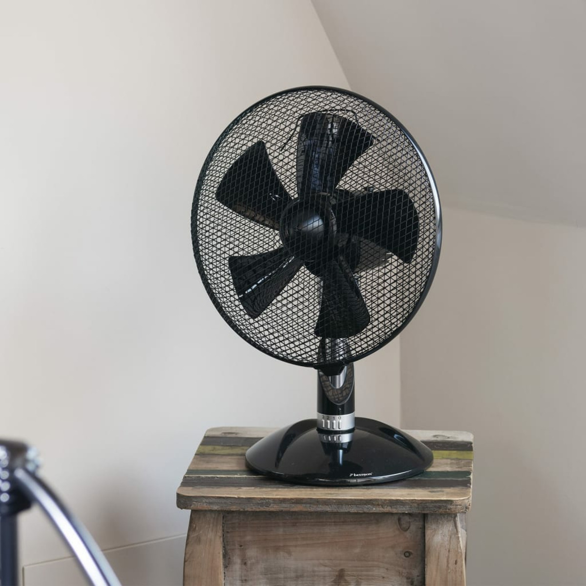 Ventilatore da tavolo nero su uno sgabello di legno, contro un muro chiaro.