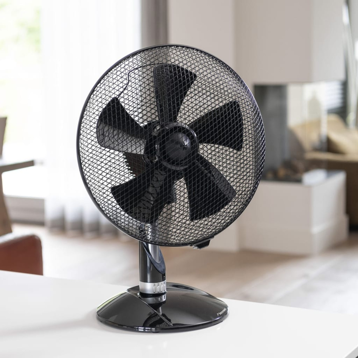 Un ventilatore da scrivania nero con una griglia a rete, su un tavolo bianco, con sfondo sfocato.
