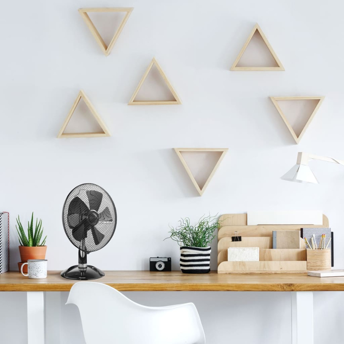Scrivania con ventilatore, piante, mensole e una sedia bianca. Scrivania in legno chiaro, muro bianco.