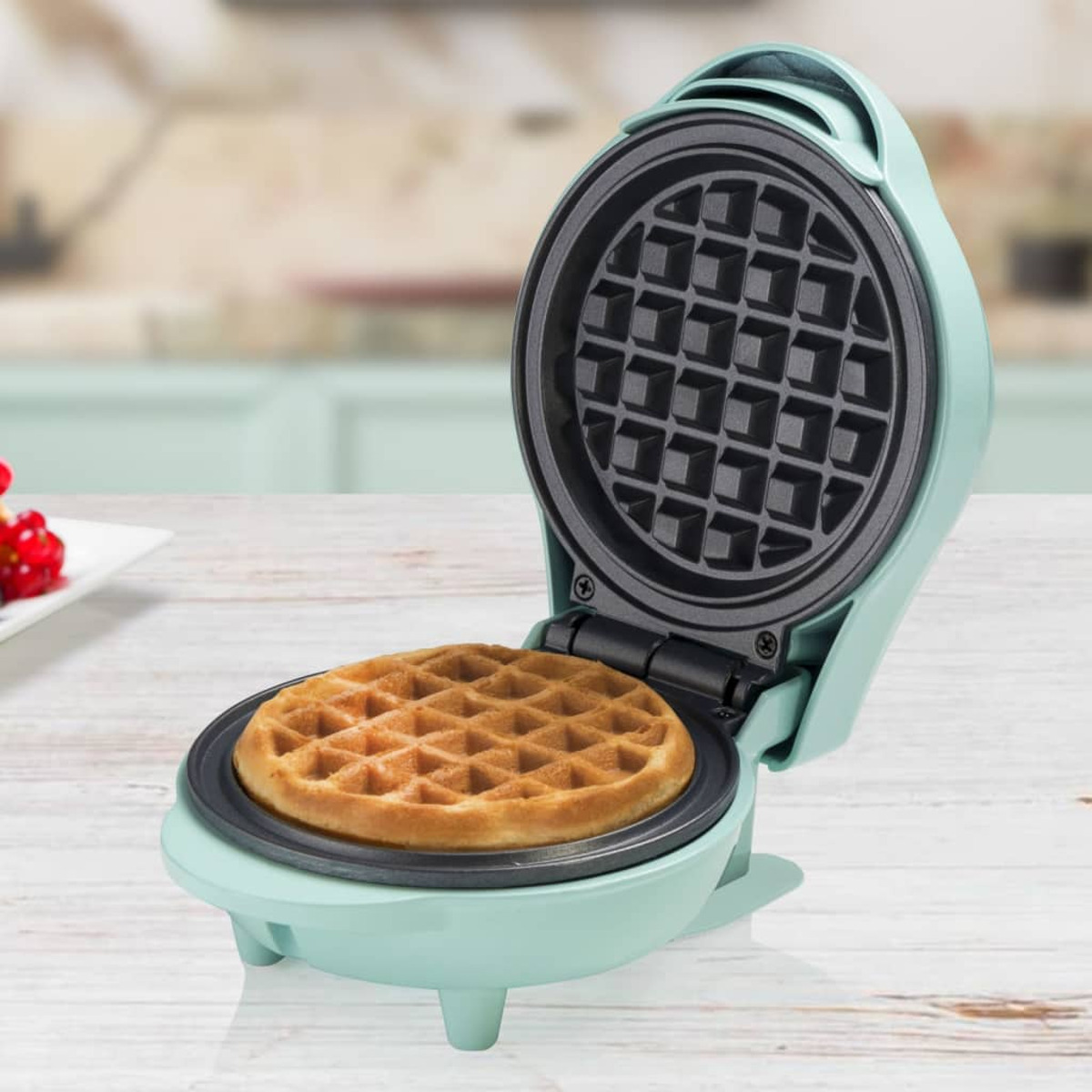 Ein Waffeleisen mit einer Waffel darin. Die Maschine ist türkis, auf einem Holztisch.
