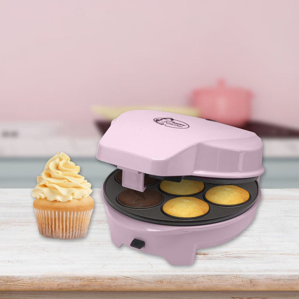 Ein pinkfarbener Cupcake-Maker mit gebackenen Cupcakes und einem vereisten Cupcake. Der Hintergrund ist eine verschwommene Küche.