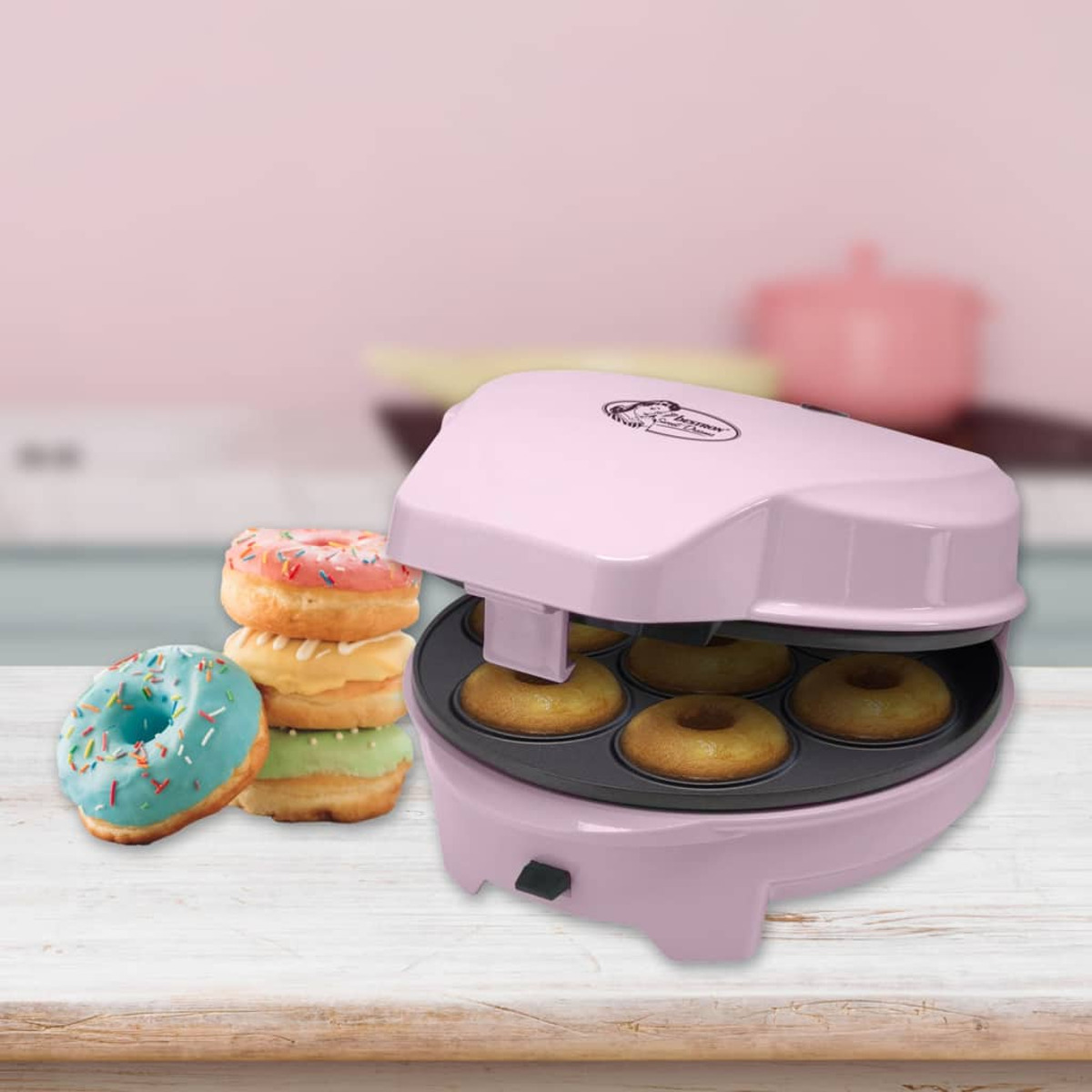 Ein rosa Donut-Maker mit Donuts daneben. Donuts sind glasiert, mit Streuseln, auf einem Holztisch.