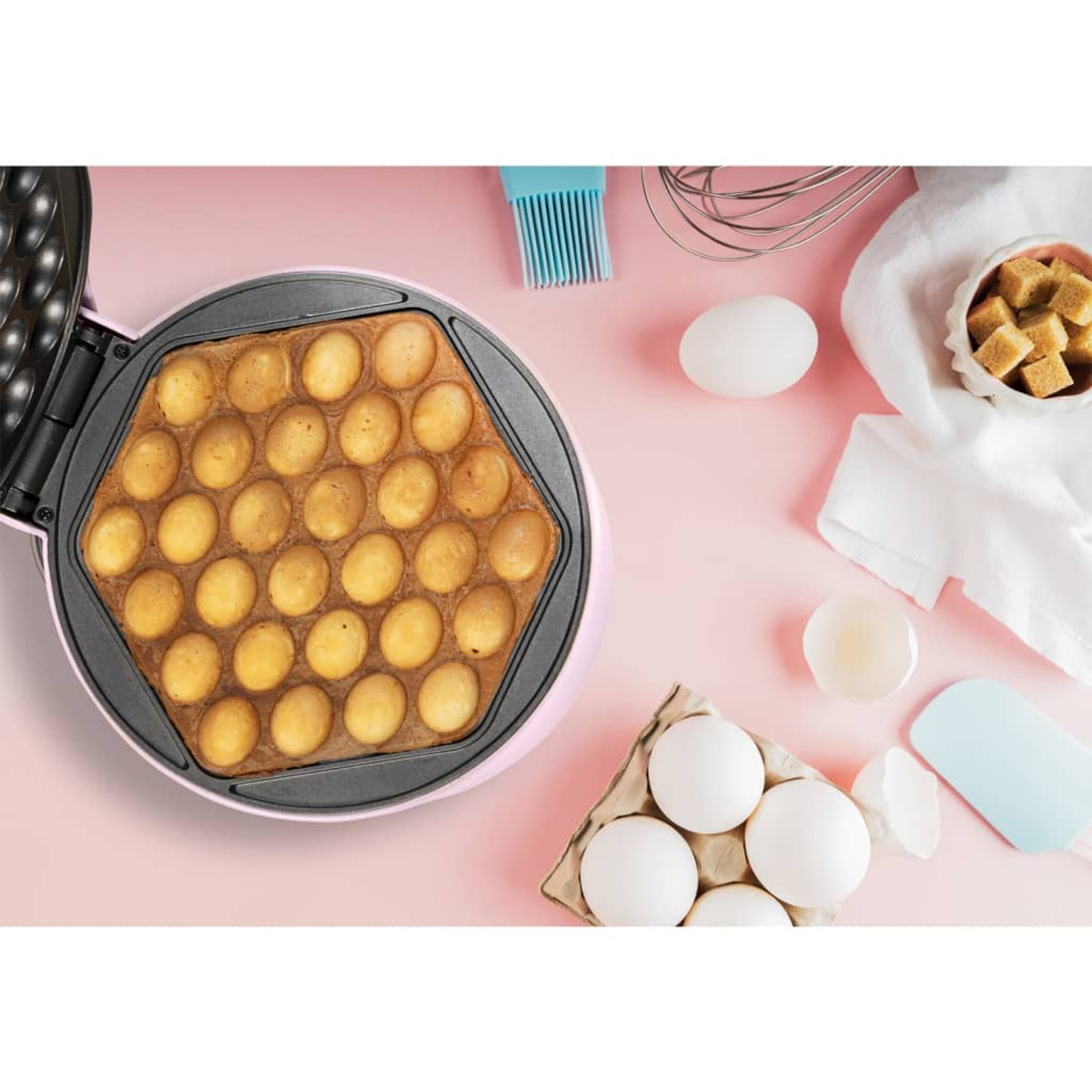 Una macchina per waffle aperta, piena di waffle cotti. Ci sono uova, zollette di zucchero e un pennello.