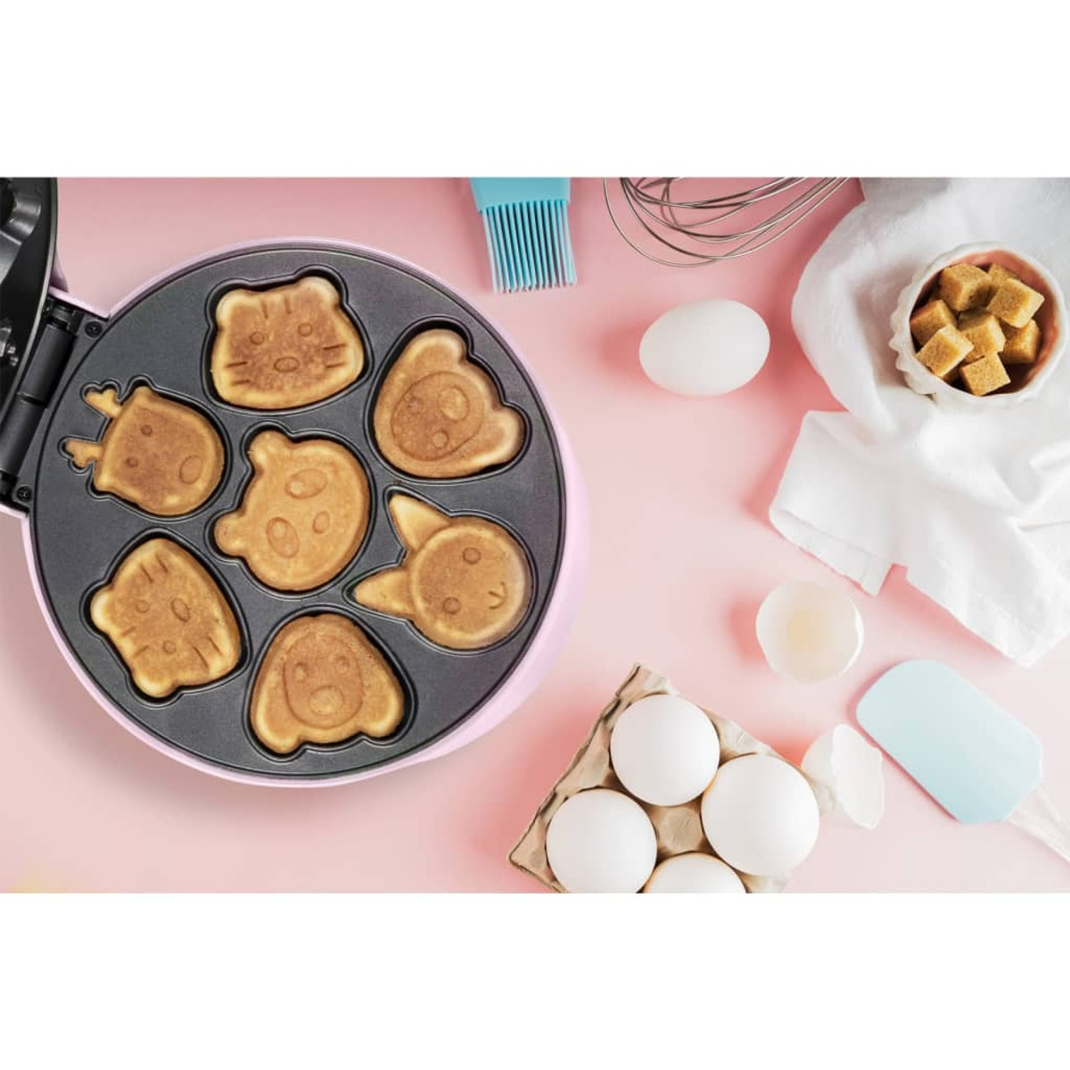 Mini macchina per pancake con forme di animali. Uova, frusta e una ciotola di zollette di zucchero sul lato.