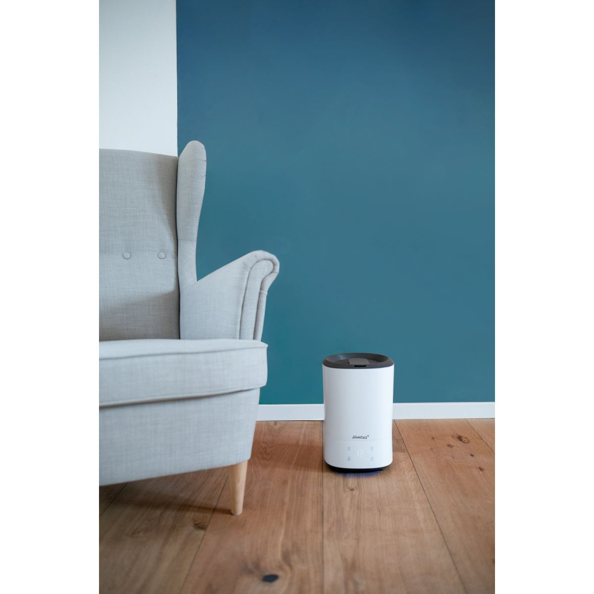 Un humidificador de aire blanco en un suelo de madera junto a una silla gris y una pared verde azulado.