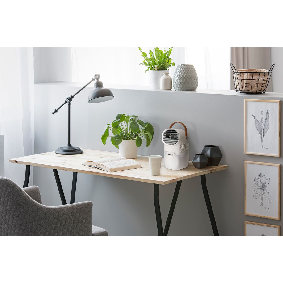 Schreibtisch mit Lampe, Pflanze, Buch und Ventilator. Fenster und gerahmte Bilder im Hintergrund.