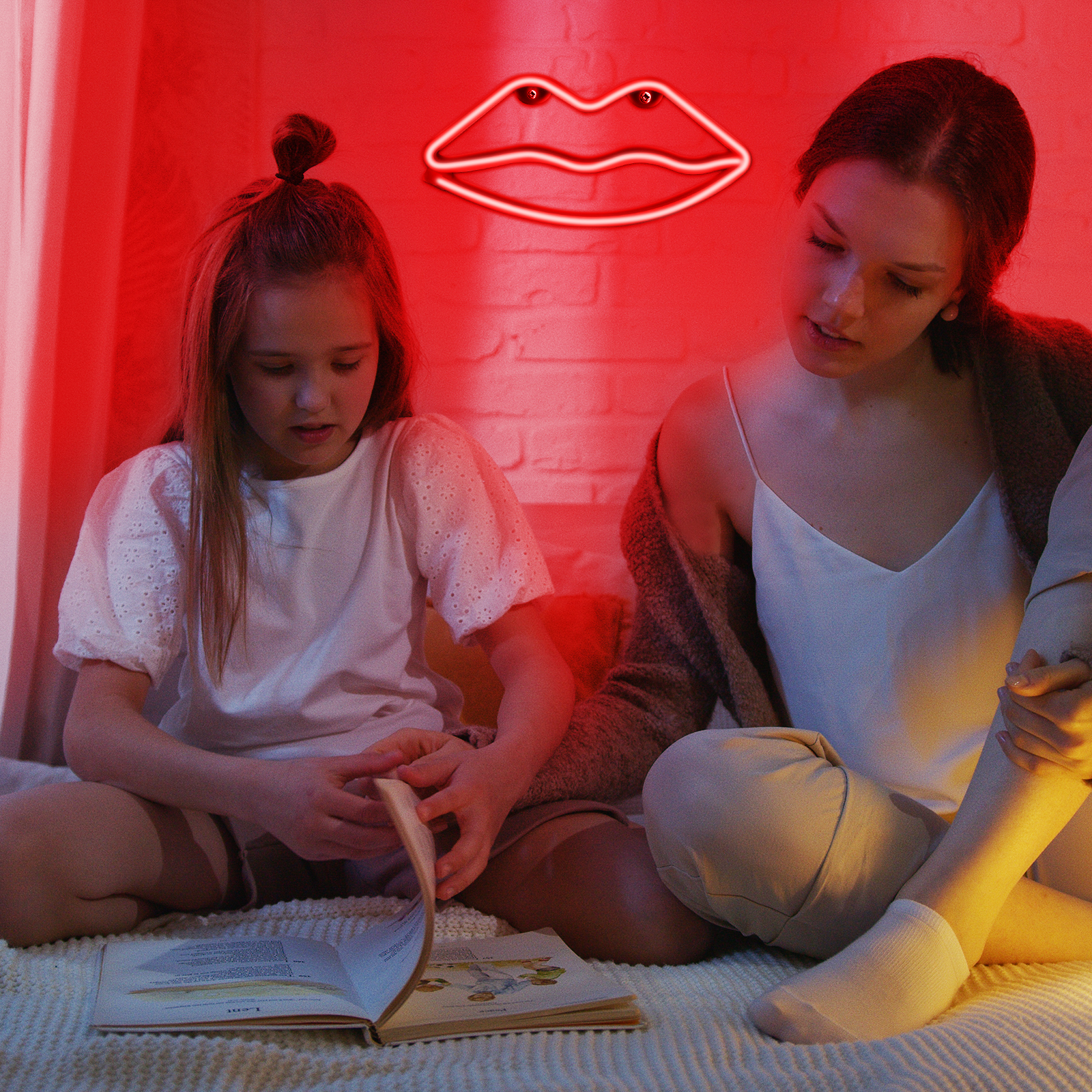 Una niña y una mujer mirando un libro. Un letrero de labios de neón está en la pared. La luz es roja.