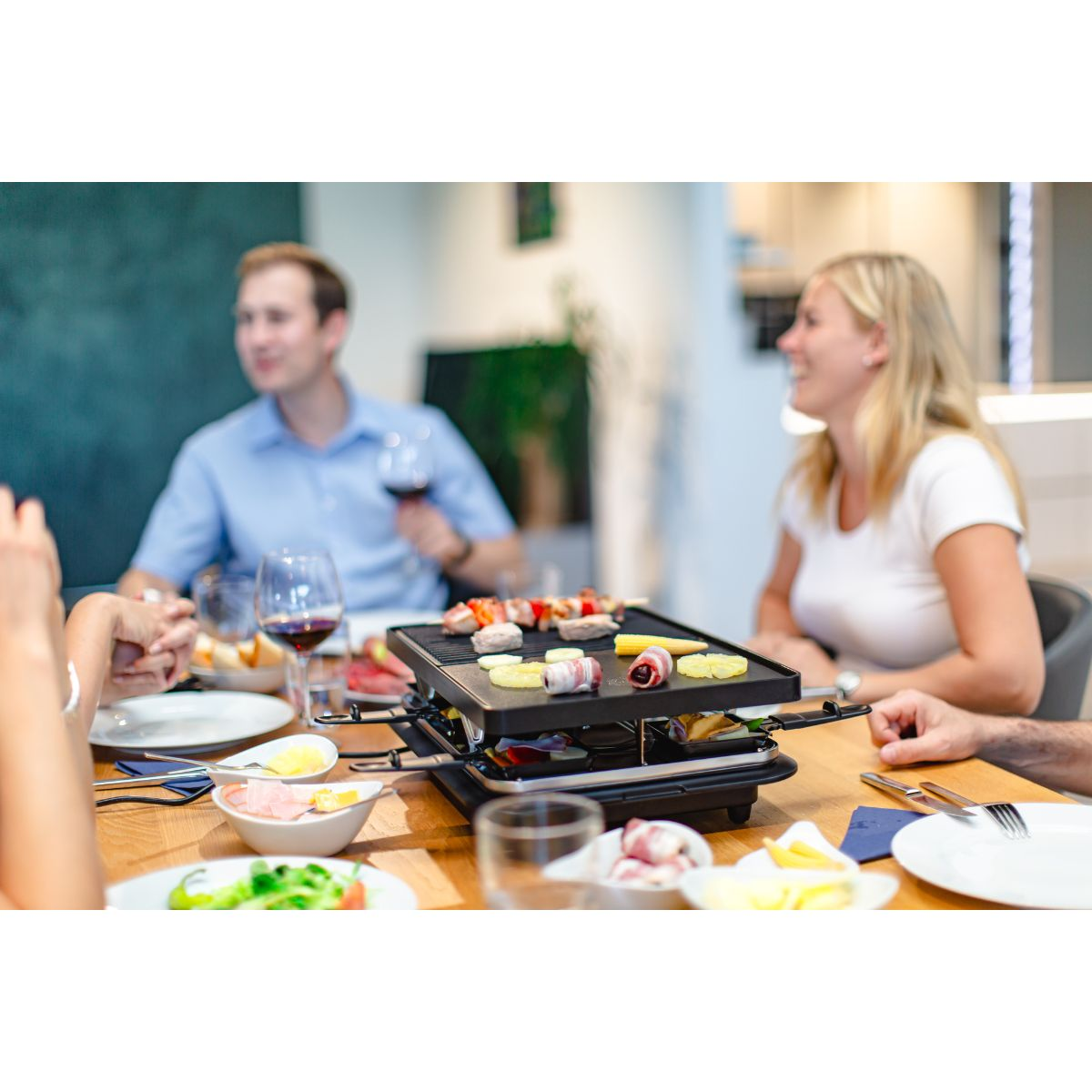 Eine Gruppe von Personen um einen Tisch mit einem Raclette-Grill, Essen wird zubereitet, Weingläser.