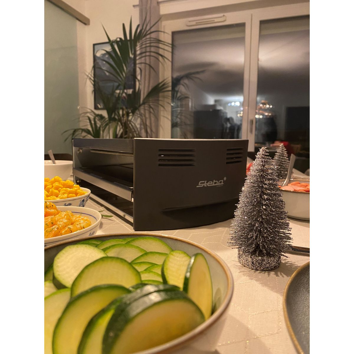 Ein Tisch mit Steba-Grill, Essen und einem Weihnachtsbaum. Die Beleuchtung ist schwach.