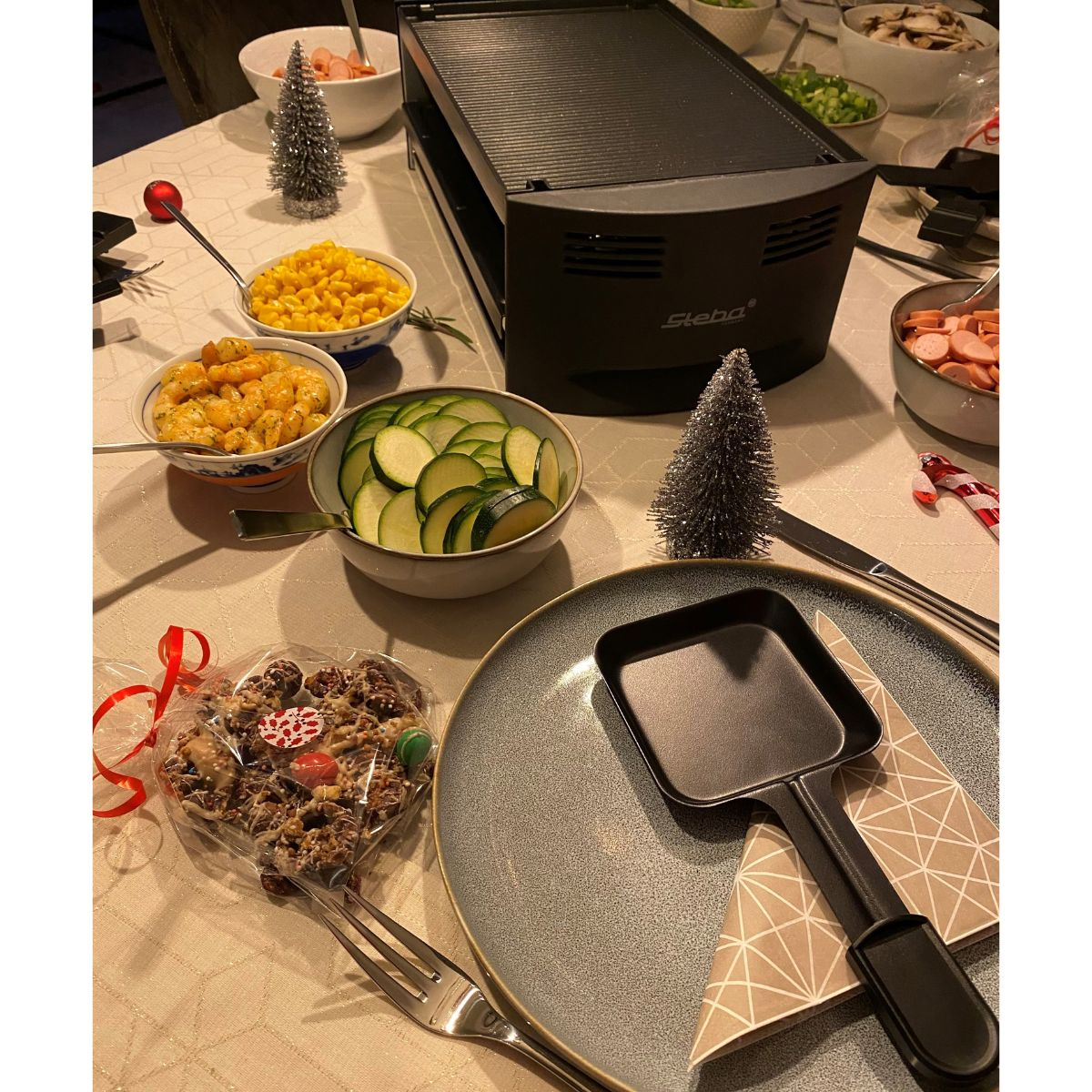 Ein Tisch mit Essen und einem Raclette-Grill. Enthält Mais, Zucchini und eine herzförmige Süßigkeit.