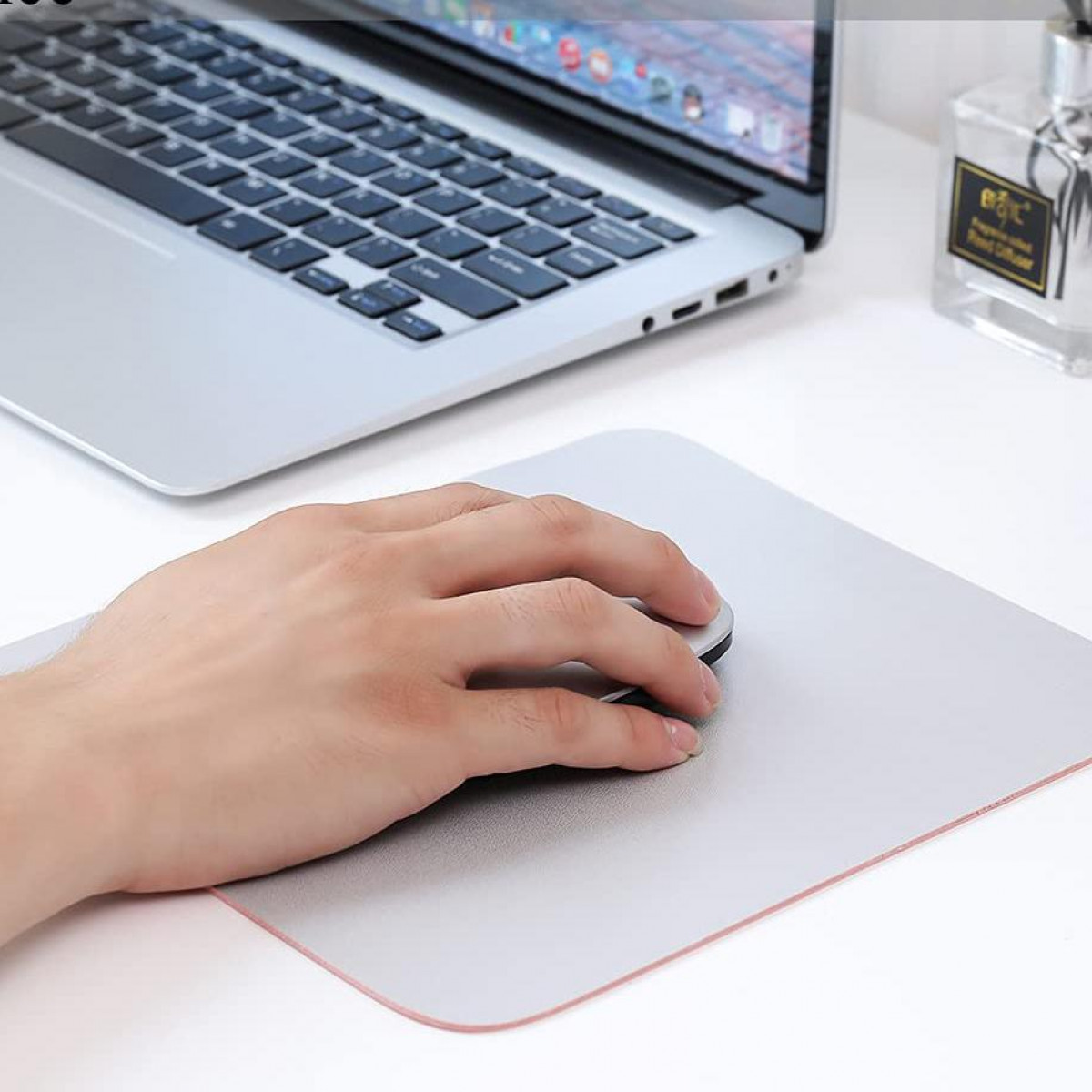 La main d'une personne utilisant une souris sur un tapis de souris. Un ordinateur portable est en arrière-plan, ainsi qu'un flacon de parfum.
