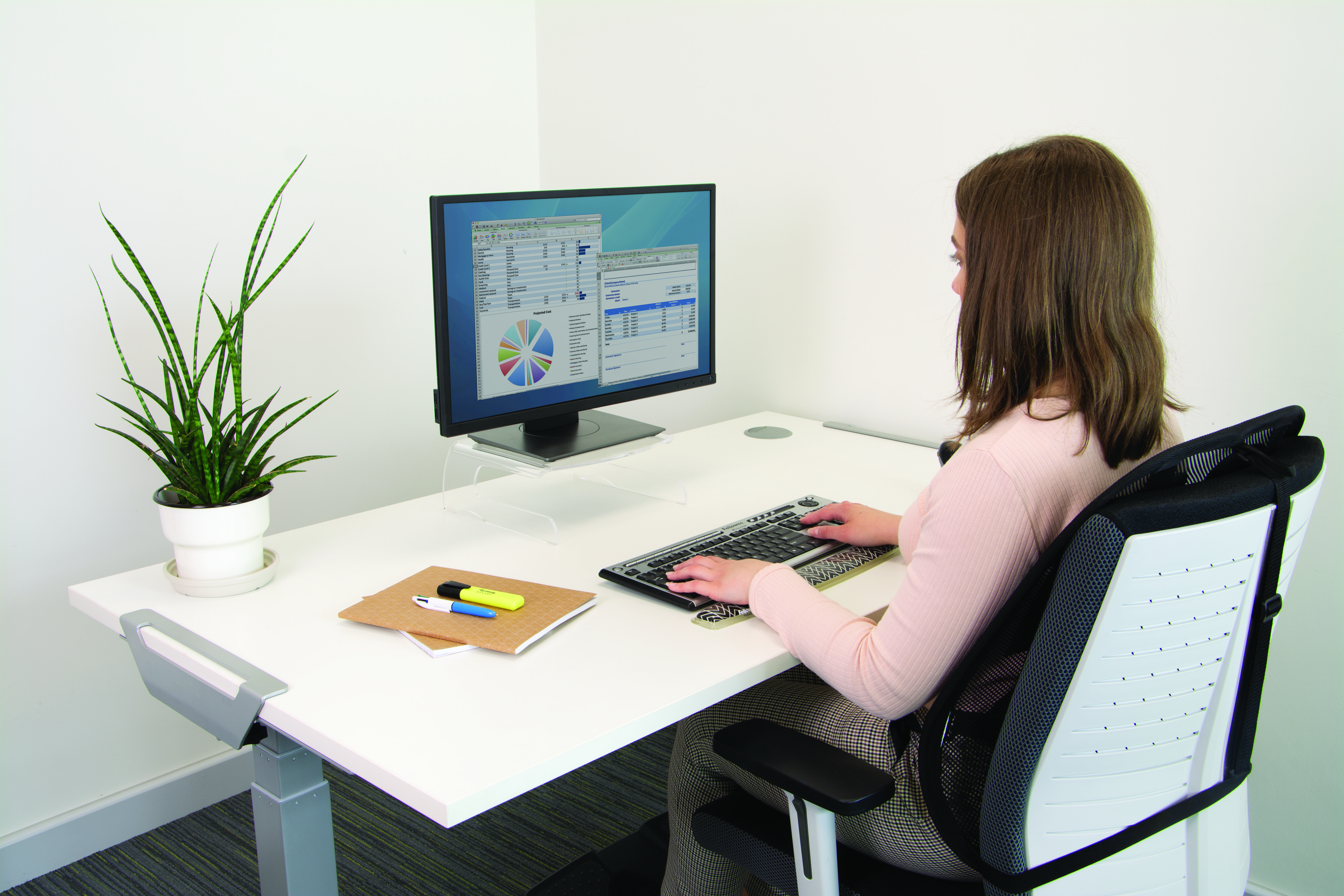 Frau tippt auf einer Tastatur an einem weißen Schreibtisch mit Monitor und Pflanze.