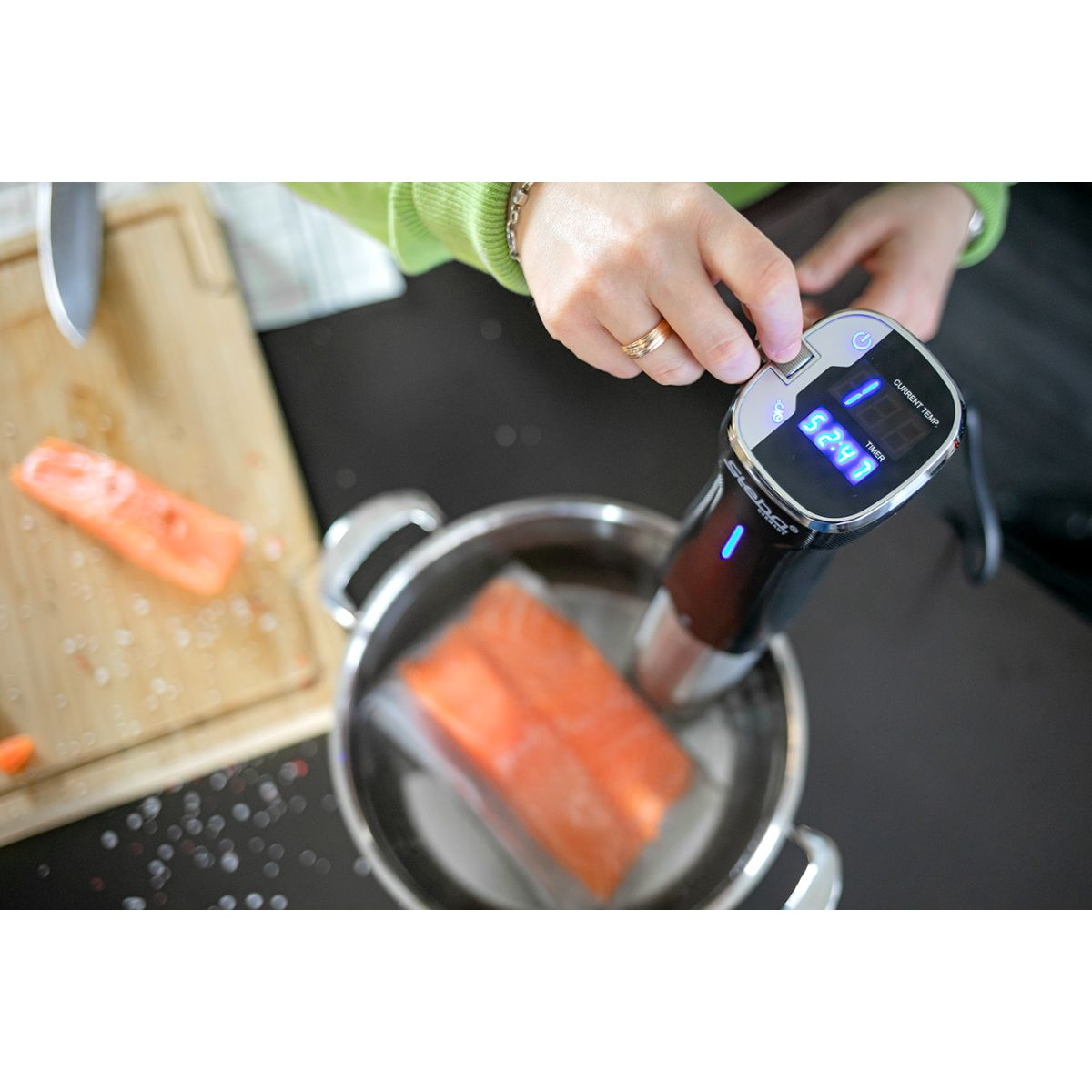 Person benutzt einen Sous-Vide-Zirkulator in einem Topf mit Lachsfilets. Das Gerät zeigt die Temperatur.