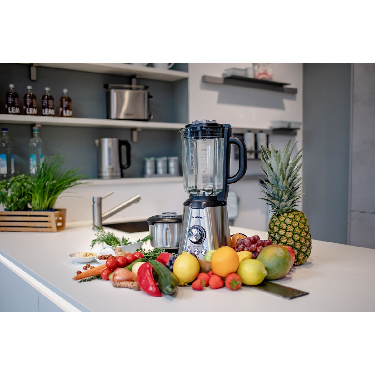 Mixer mit Obst auf einer Arbeitsplatte. Es gibt eine Ananas, Zitronen, Erdbeeren und anderes Obst.