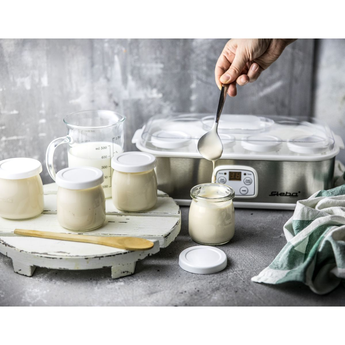 Joghurt Herstellungsprozess. Hand gießt Joghurt von einem Löffel in ein Glas. Andere Gläser und Zutaten.