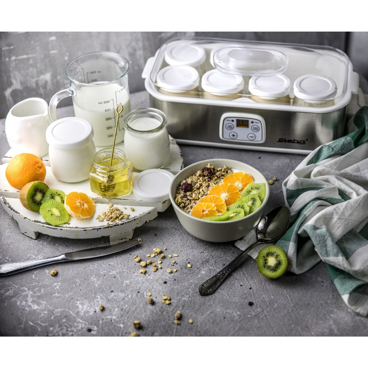 Joghurtbereiter mit Joghurtgläsern, Obst und Müsli in einer Schüssel.