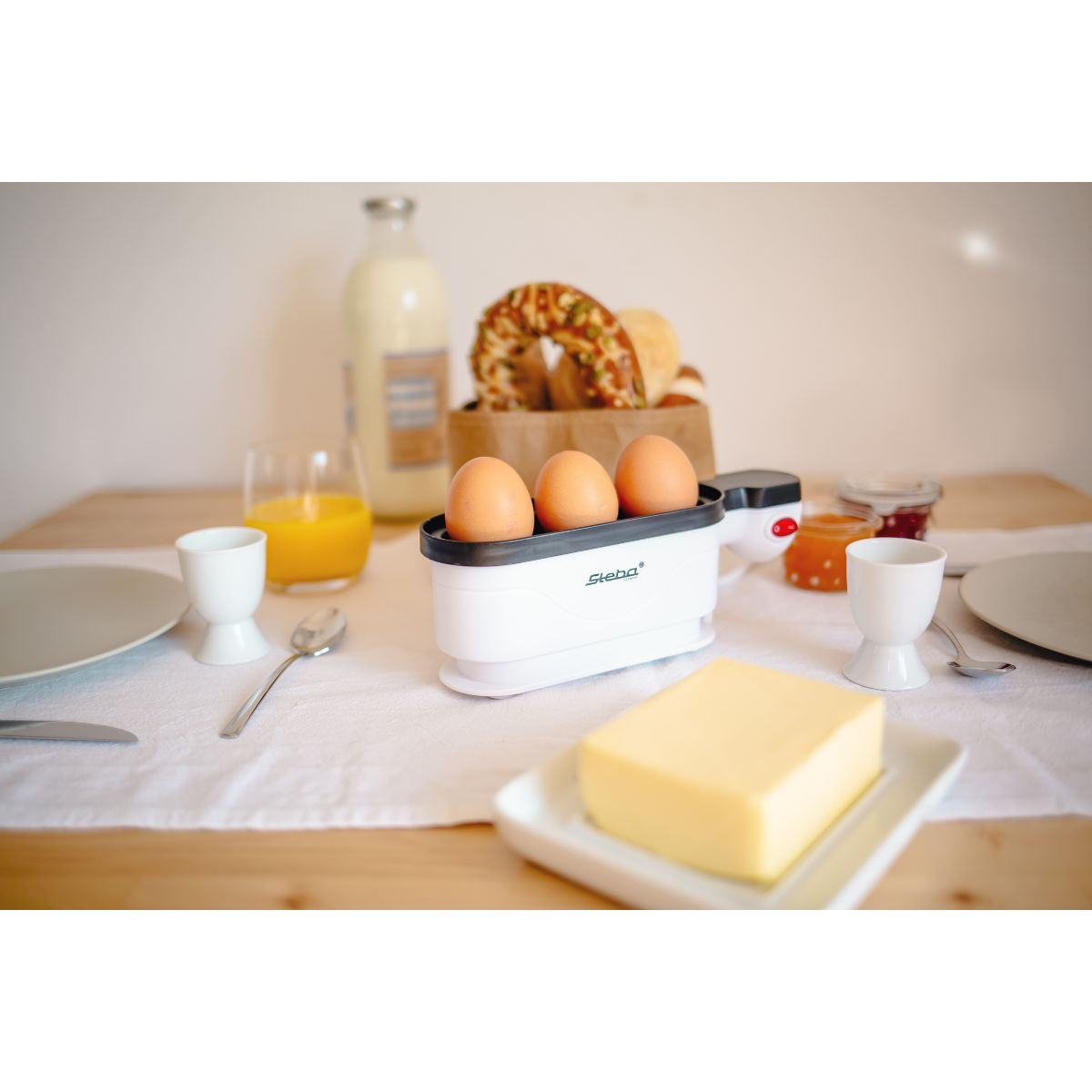 Jajka w urządzeniu do gotowania jajek na stole z jedzeniem, jak masło, sok i chleb.