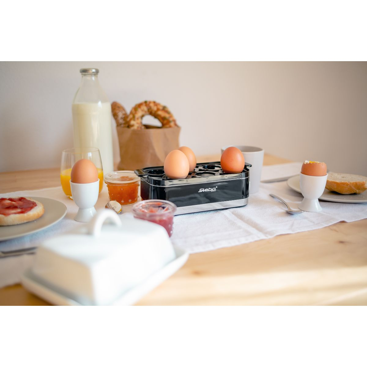 Frühstückstisch mit Eiern, Brot, Marmelade und einer Milchflasche. Schwarzer Eierkocher sichtbar.