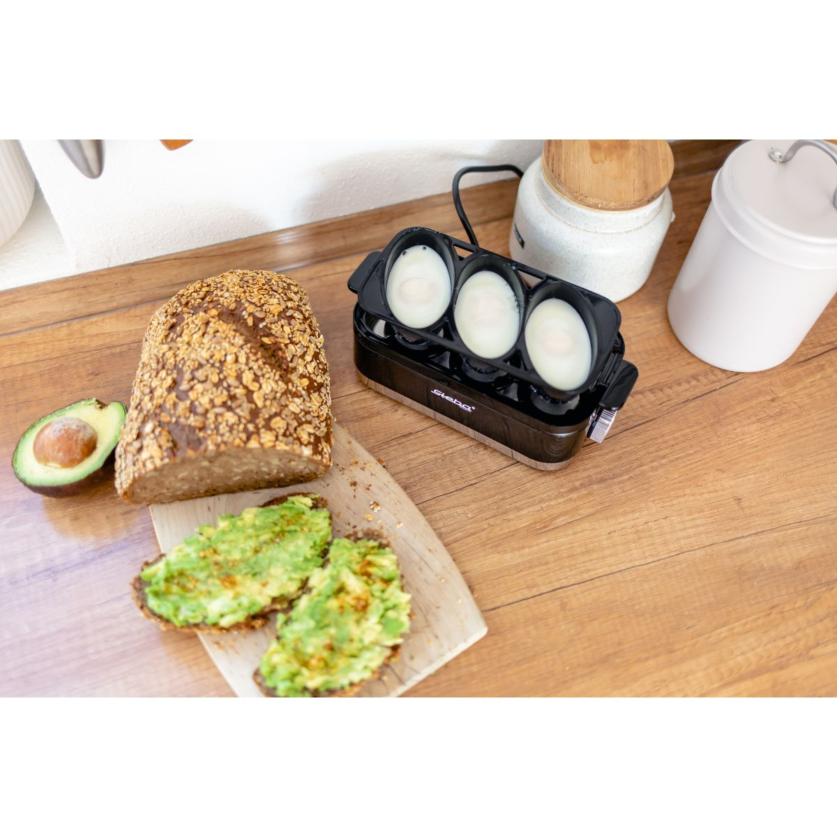 Ein Eierkocher mit drei Eiern, Brot, Avocado und Toast auf einer Holzoberfläche.