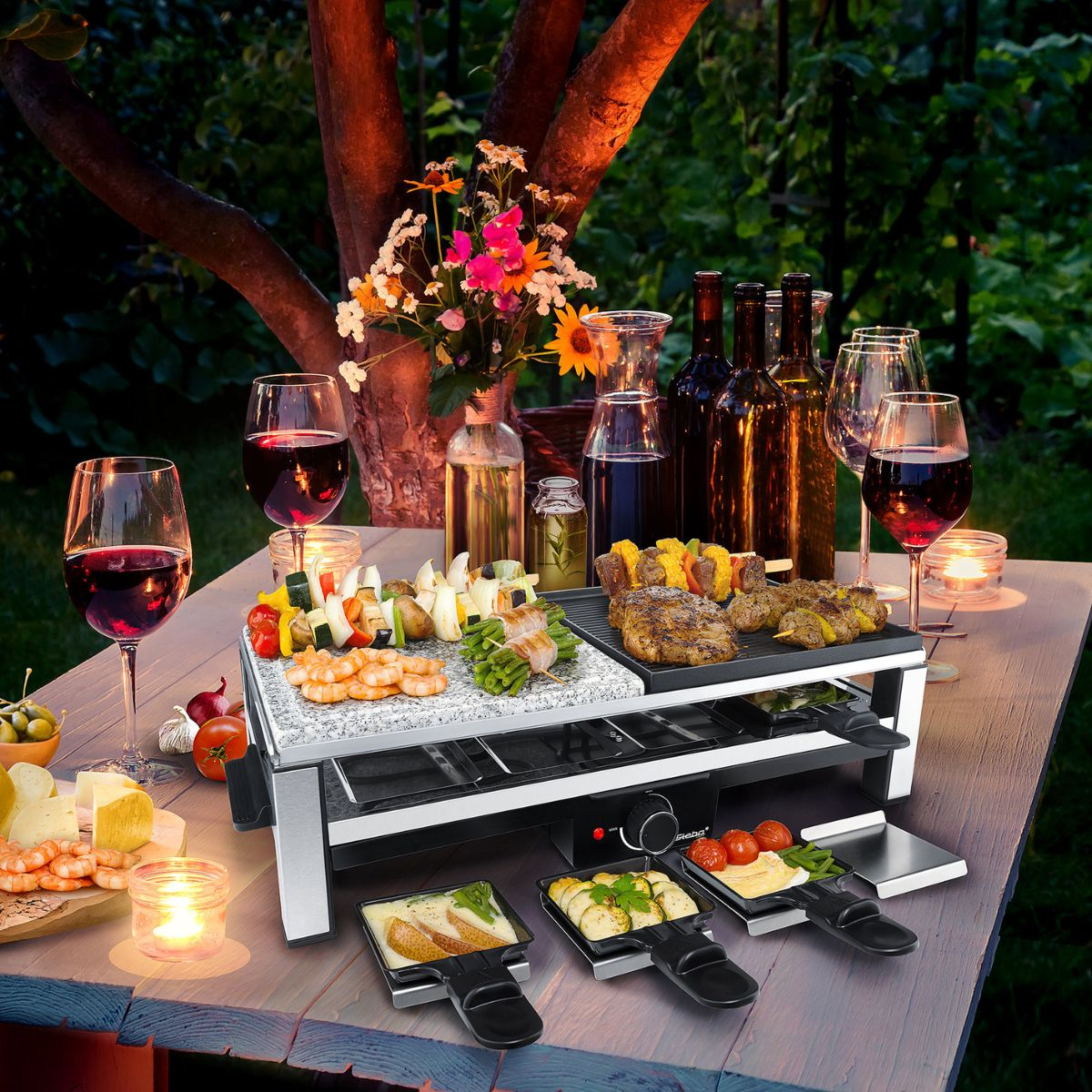 Ein Tisch mit einem Raclette-Grill, Lebensmitteln und Weingläsern im Freien bei Dämmerung.