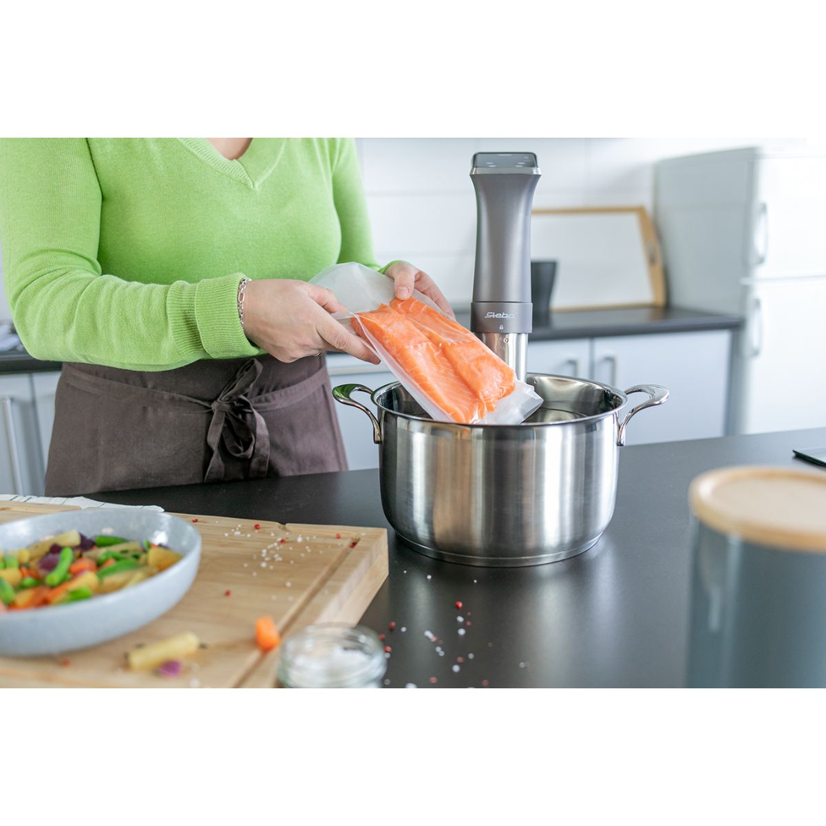 Eine Frau in einem grünen Hemd legt Lachs in einen Topf. Eine Sous-Vide-Maschine ist im Topf.