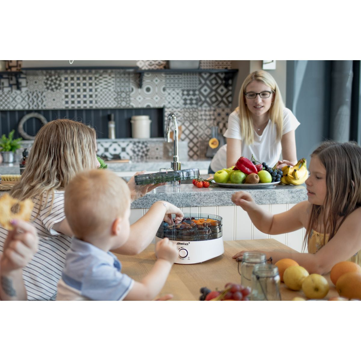 Une famille est réunie autour d'un déshydrateur, avec une femme en arrière-plan. Fruits et légumes.