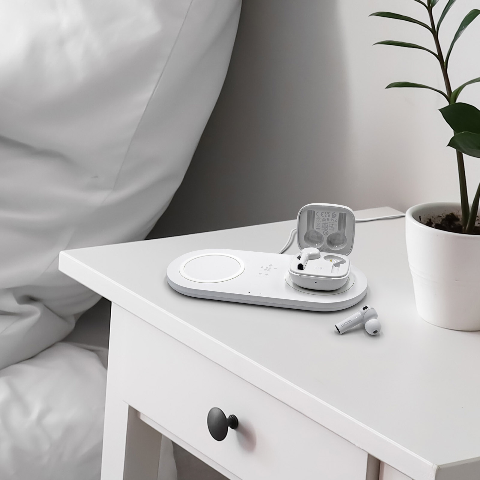 Weißer Nachttisch mit kabellosem Ladepad, Ohrhörern und Pflanze. Bett und Wand im Hintergrund.