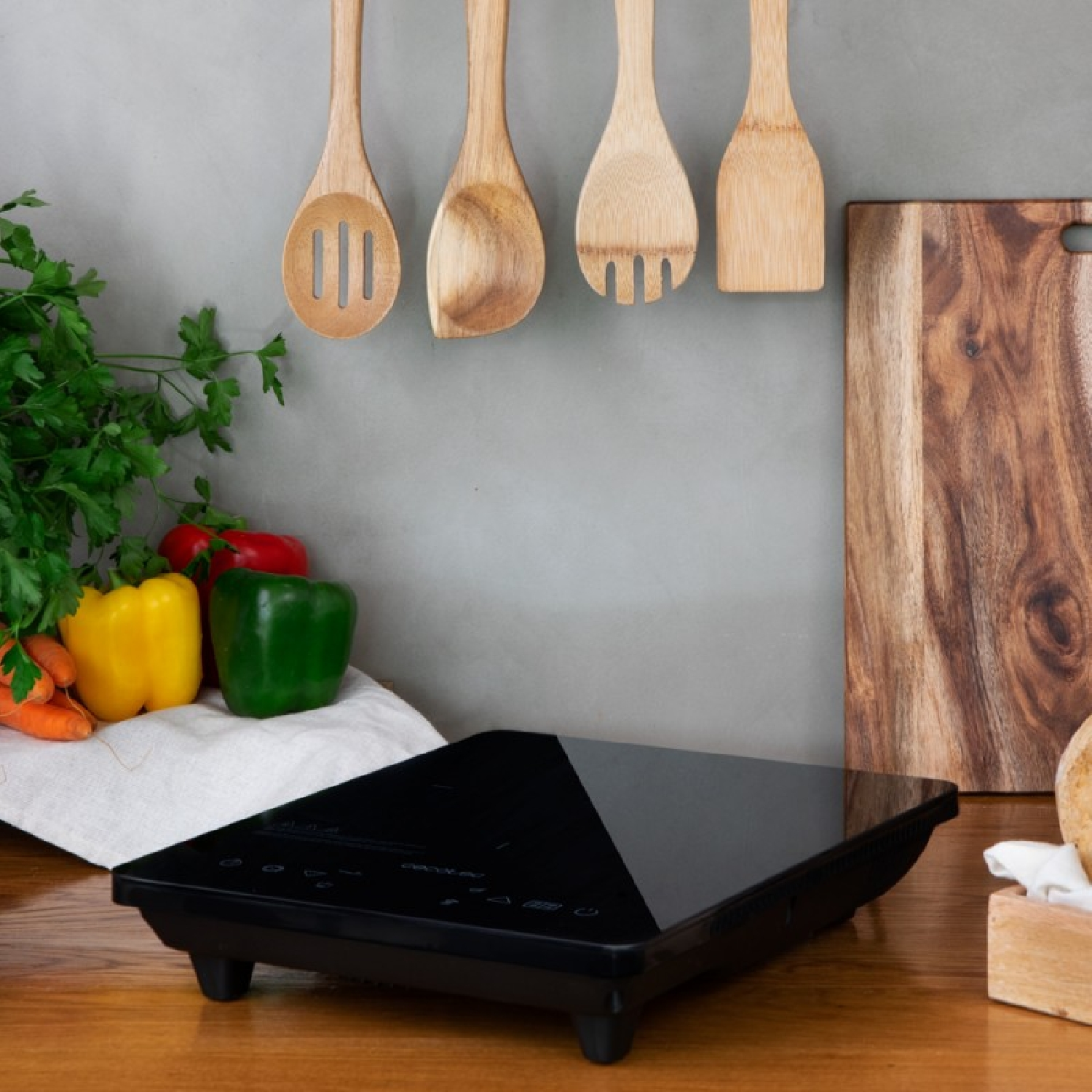 Czarna płyta indukcyjna na drewnianej powierzchni. Drewniane szpatułki i warzywa w tle.