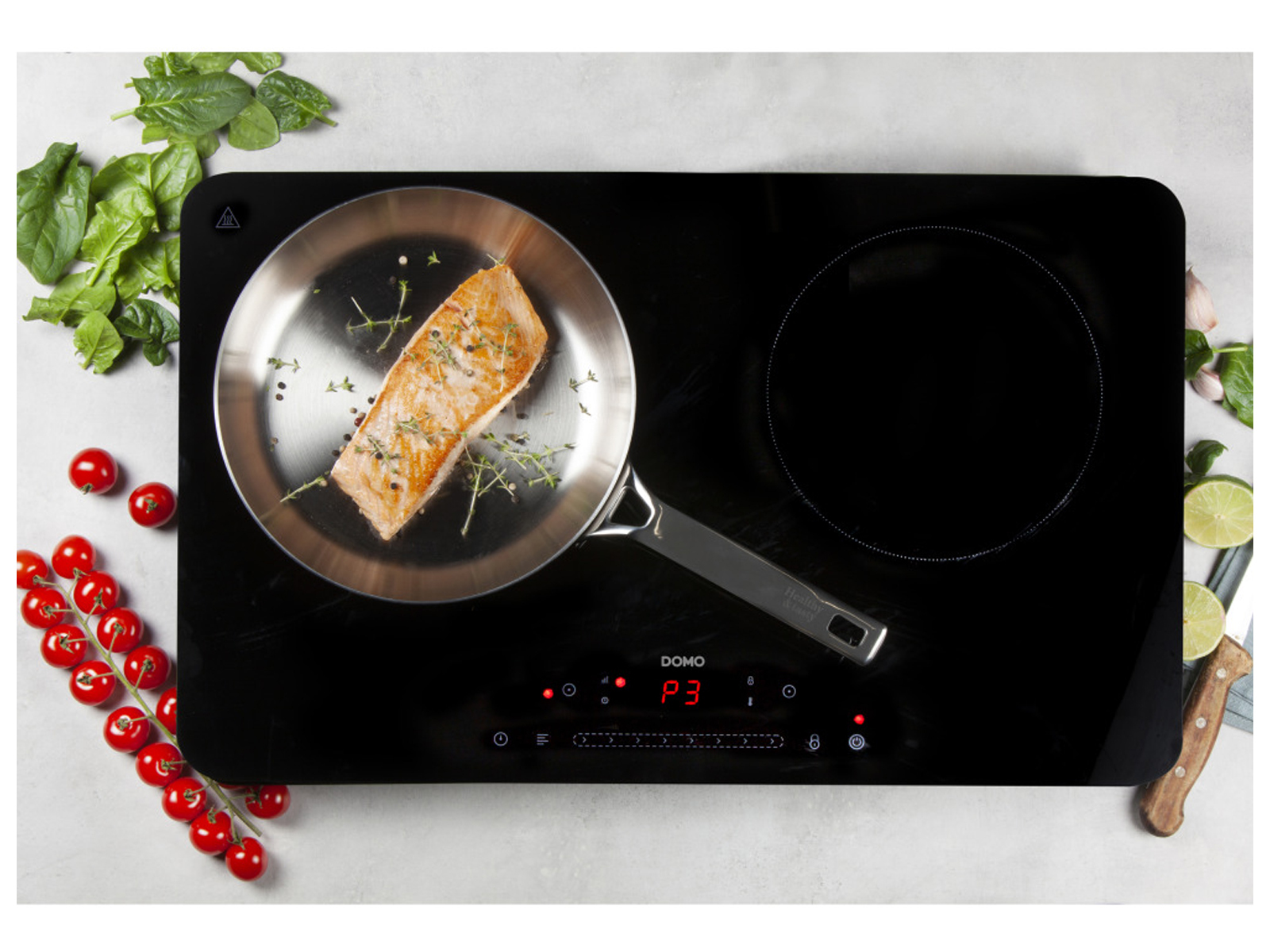 Table de cuisson à induction noire avec poêle de saumon cuit, entourée d'ingrédients.