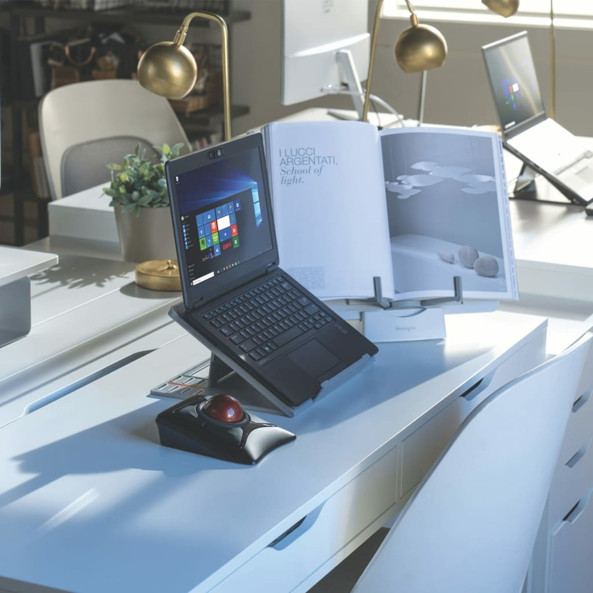 Laptop on a desk with books, a mouse, and lamps. It is a bright room.