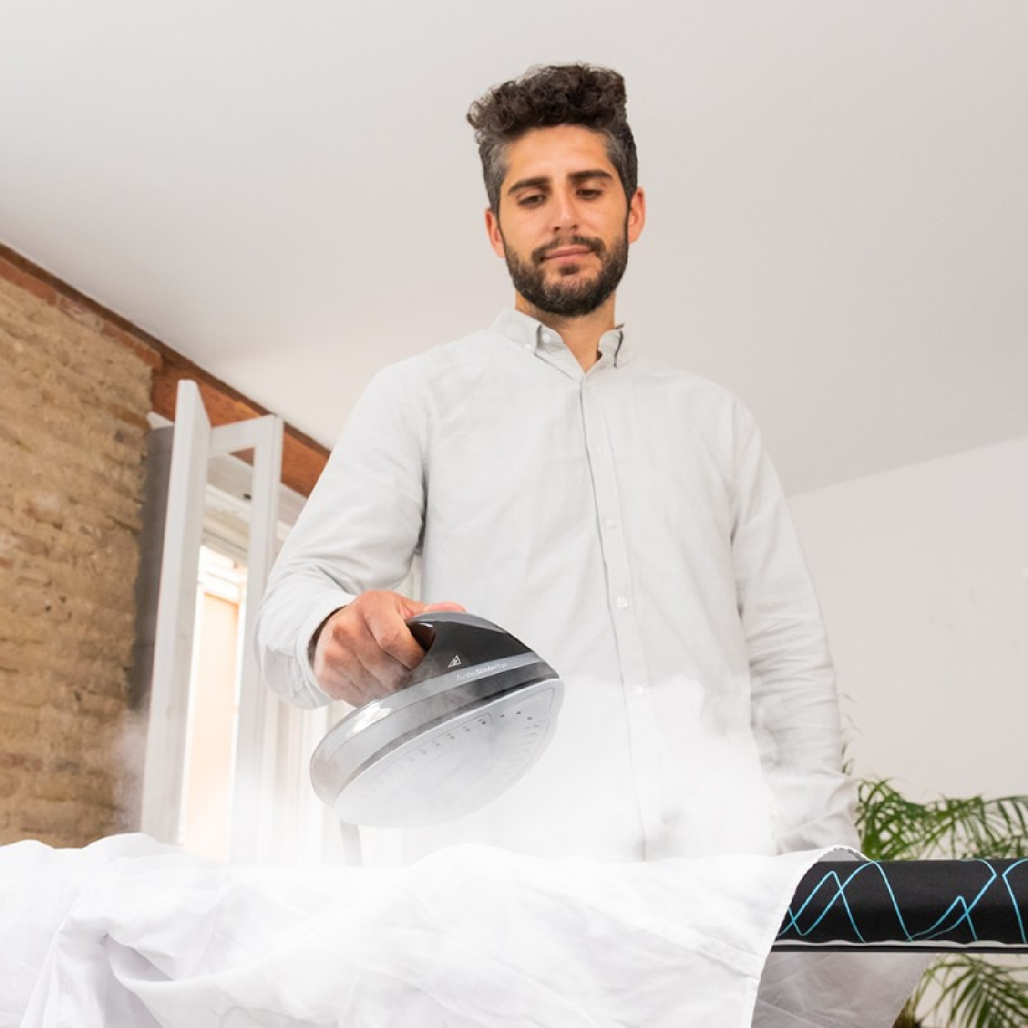Un hombre planchando una camisa blanca en una tabla de planchar, sale vapor. Fondo claro.