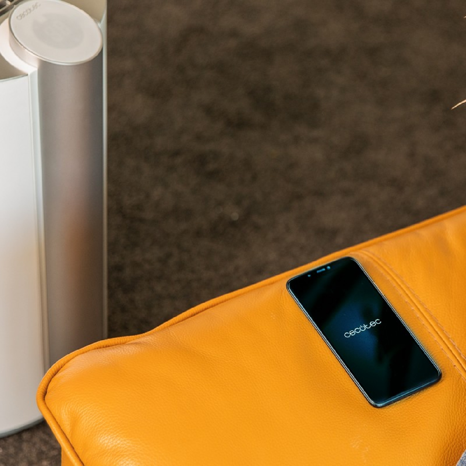 Czarny smartfon leży na pomarańczowej skórzanej kanapie obok białego oczyszczacza powietrza.