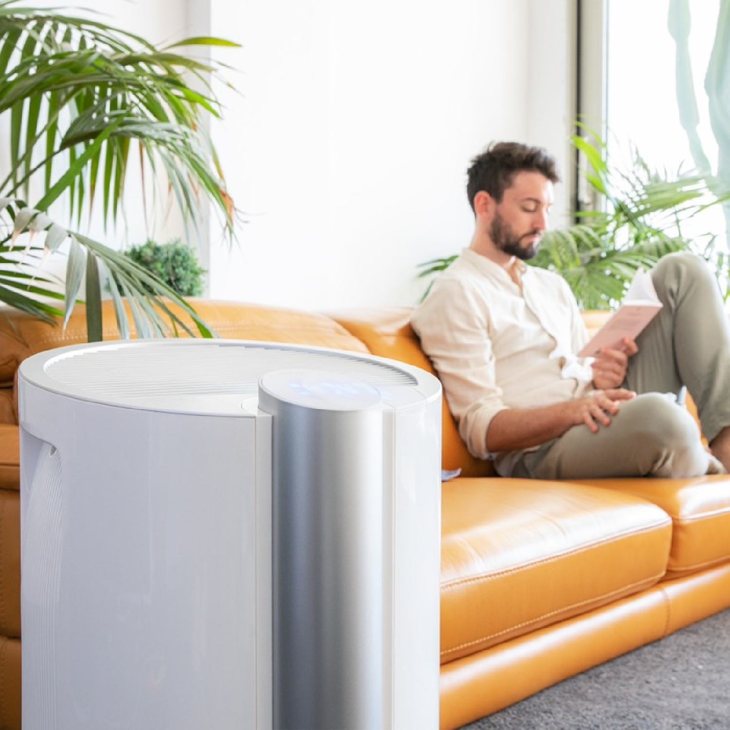 Mężczyzna czytający na sofie z białym oczyszczaczem powietrza na pierwszym planie.