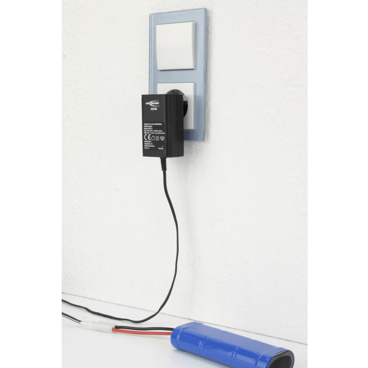 Ladegerät in einer Steckdose. Eine blaue Batterie liegt auf dem Boden. Die Wand ist weiß. Ansmann-Ladegerät.
