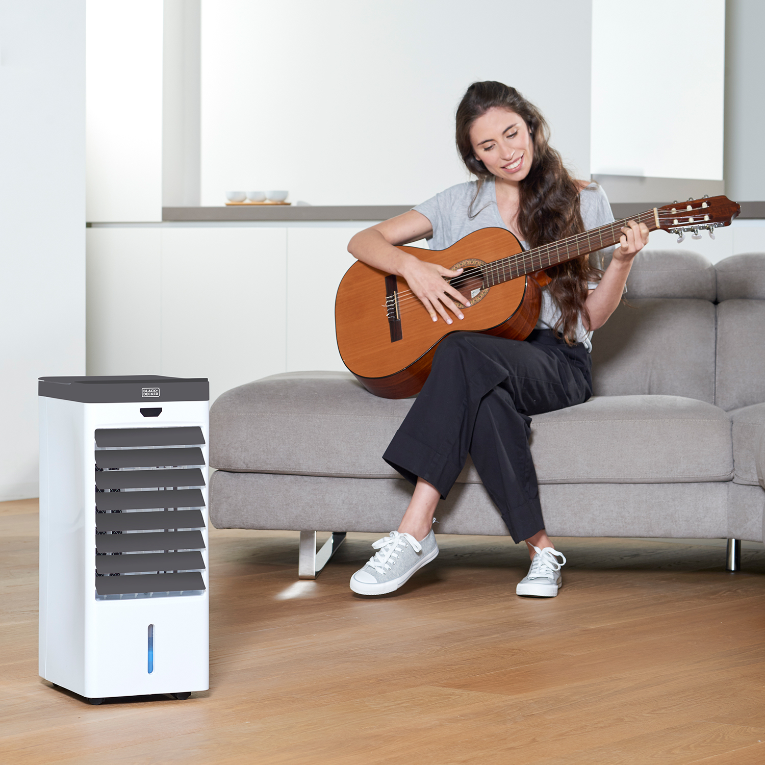 Kobieta gra na gitarze obok białego klimatyzatora na drewnianej podłodze, szara sofa w tle.