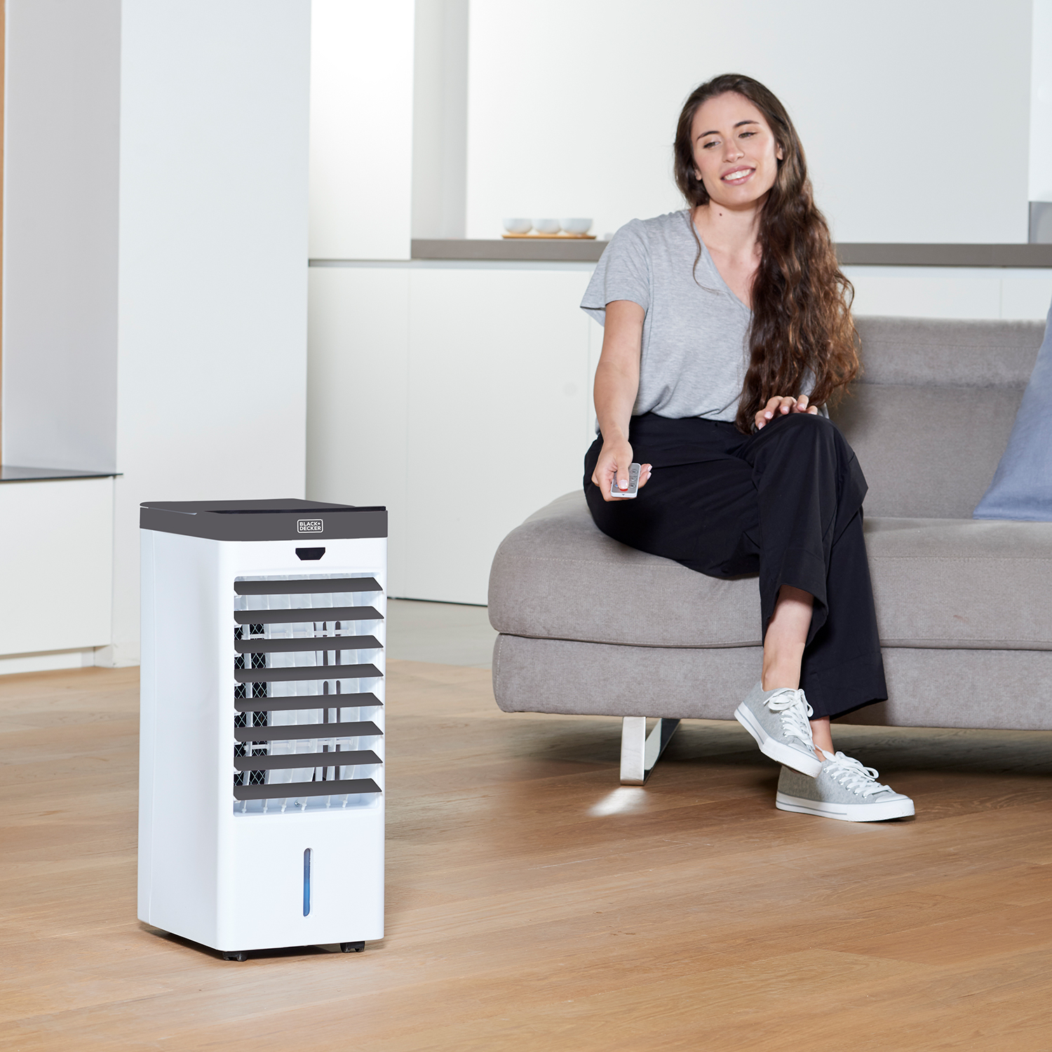 Kobieta siedzi na kanapie, używając pilota. Biała chłodziarka powietrza na pierwszym planie.