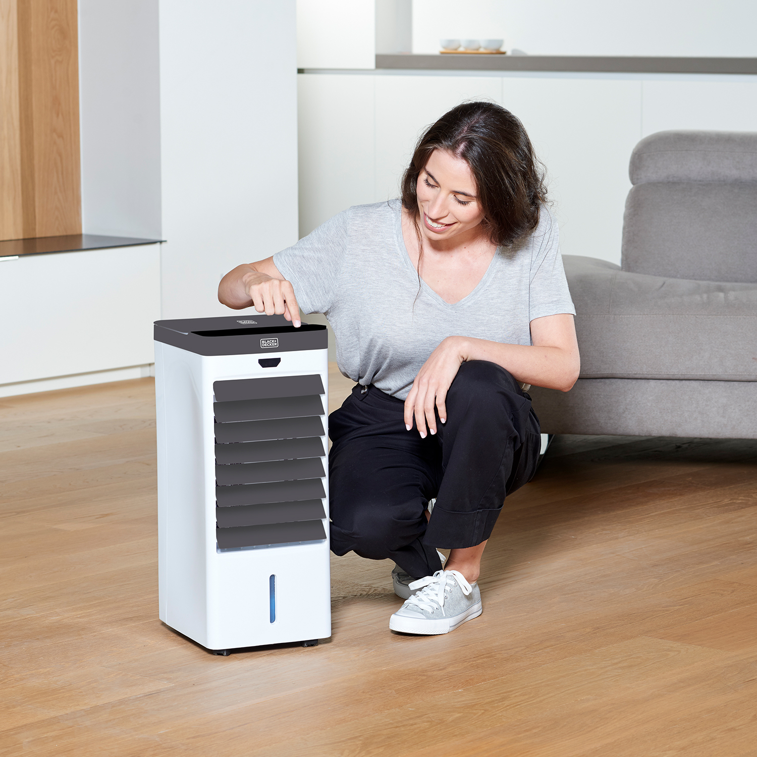 Kobieta klęcząca obok biało-czarnej chłodziarki powietrza. Drewniana podłoga, domowe otoczenie.