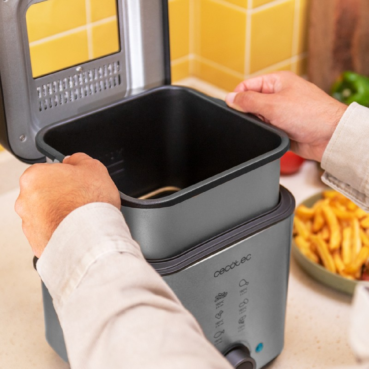 Ręce umieszczają wewnętrzny pojemnik frytkownicy Cecotec w podstawie, tło kuchni.