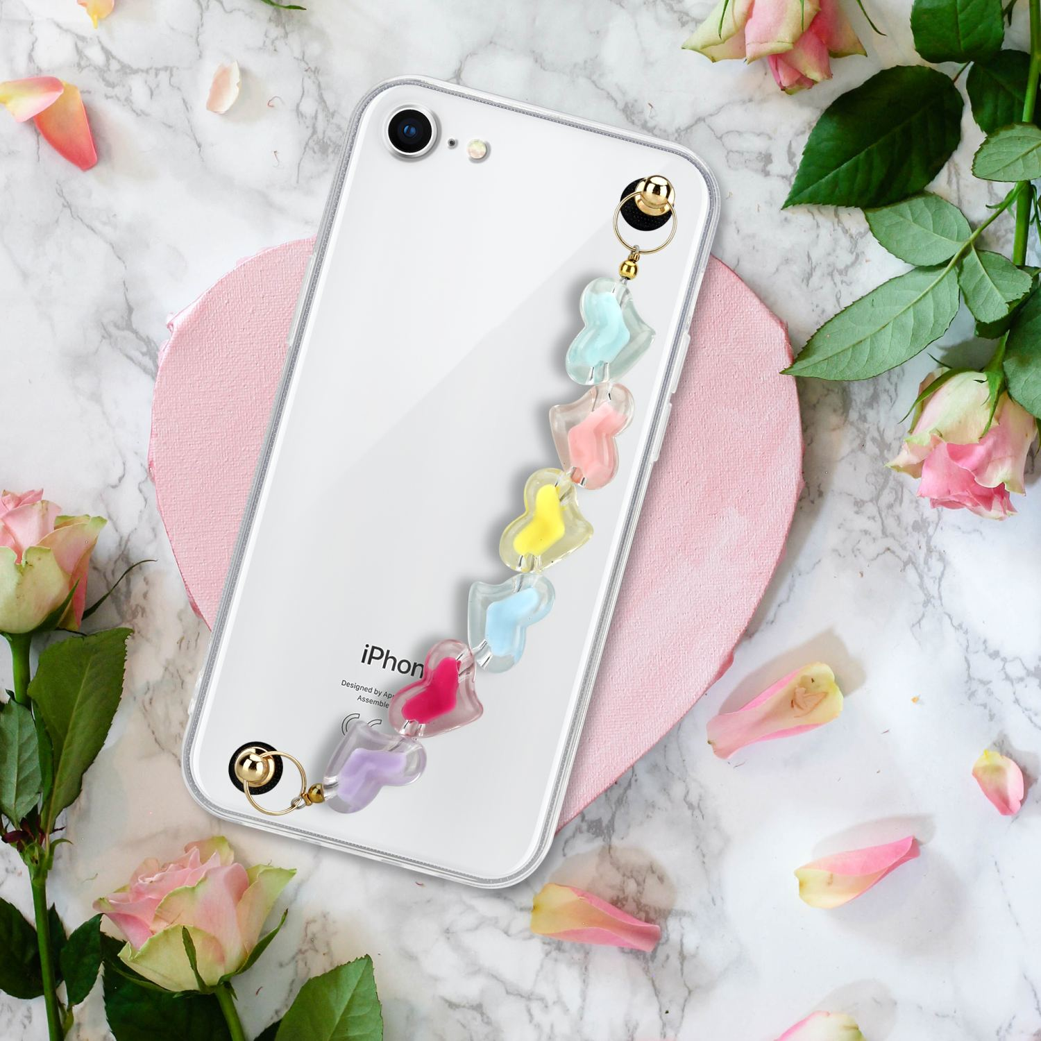 Eine transparente Handyhülle mit einem bunten Riemen mit Herzformen. Im Hintergrund sind Rosen.