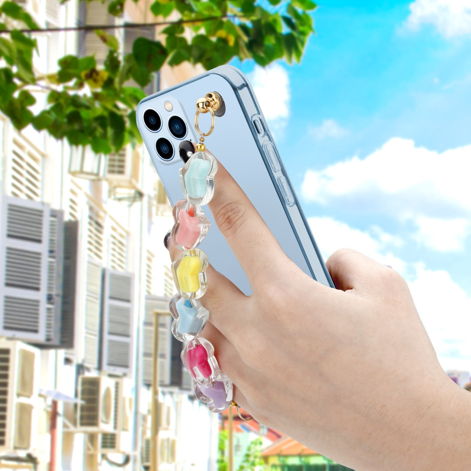 Ein klares Telefon, gehalten von einer Hand mit farbenfrohem Blumenband, vor Gebäude und blauem Himmel.