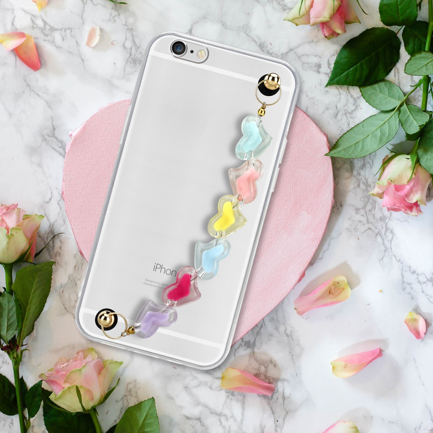 Transparente Handyhülle mit Anhänger und bunten Perlen. Rosa Herzform und Rosen im Hintergrund.