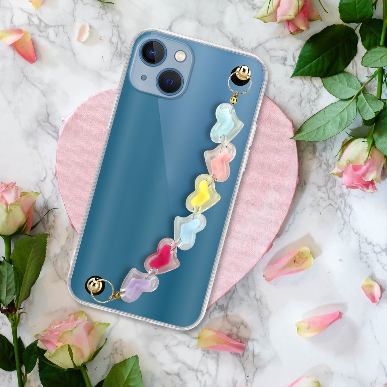 Ein Telefon in einer transparenten Hülle mit bunter herzförmiger Kette liegt zwischen Rosen und Blütenblättern auf Marmor.