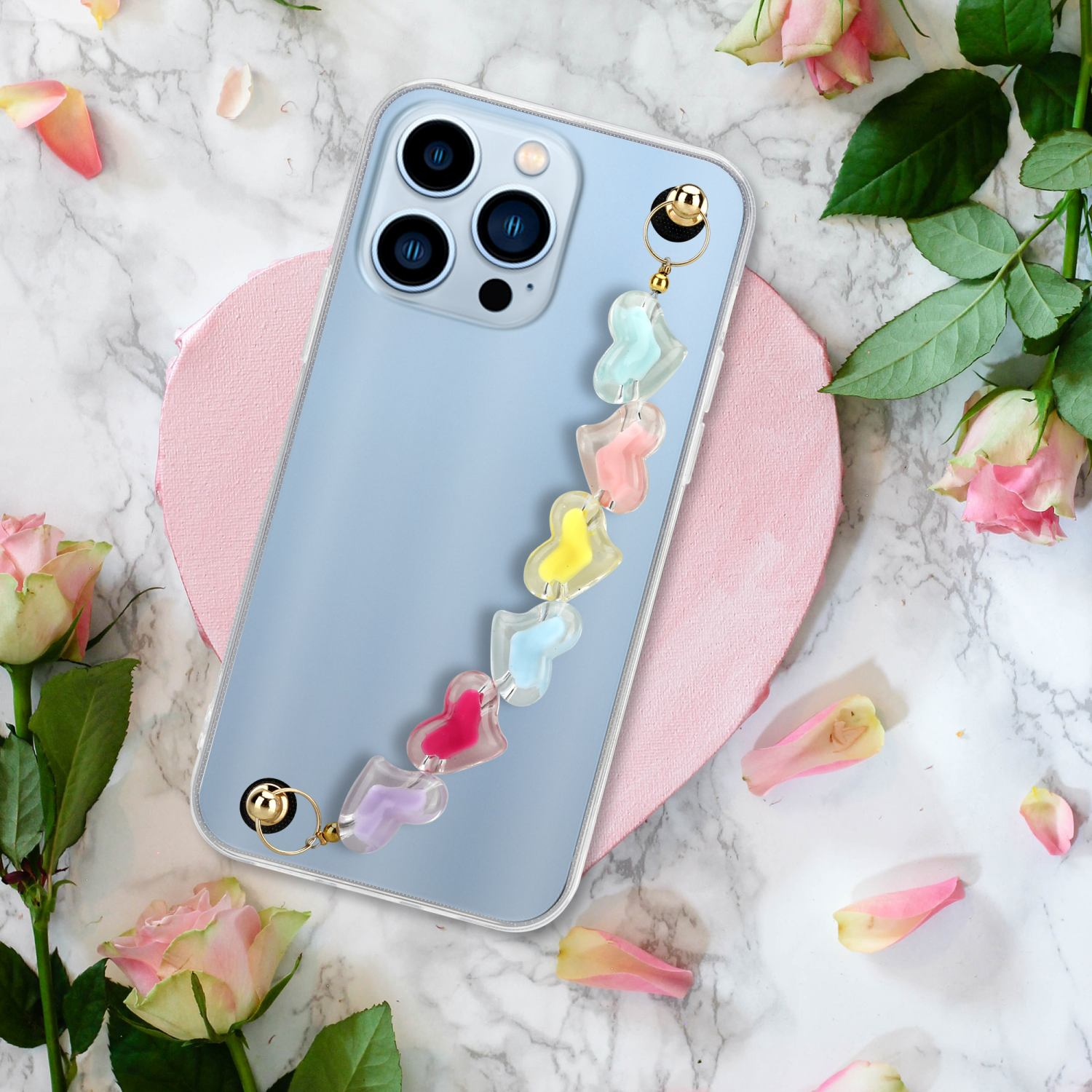 Ein Telefon mit einem bunten, herzförmigen, mit Perlen besetzten Riemen, umgeben von Rosen und Blütenblättern auf Marmor.