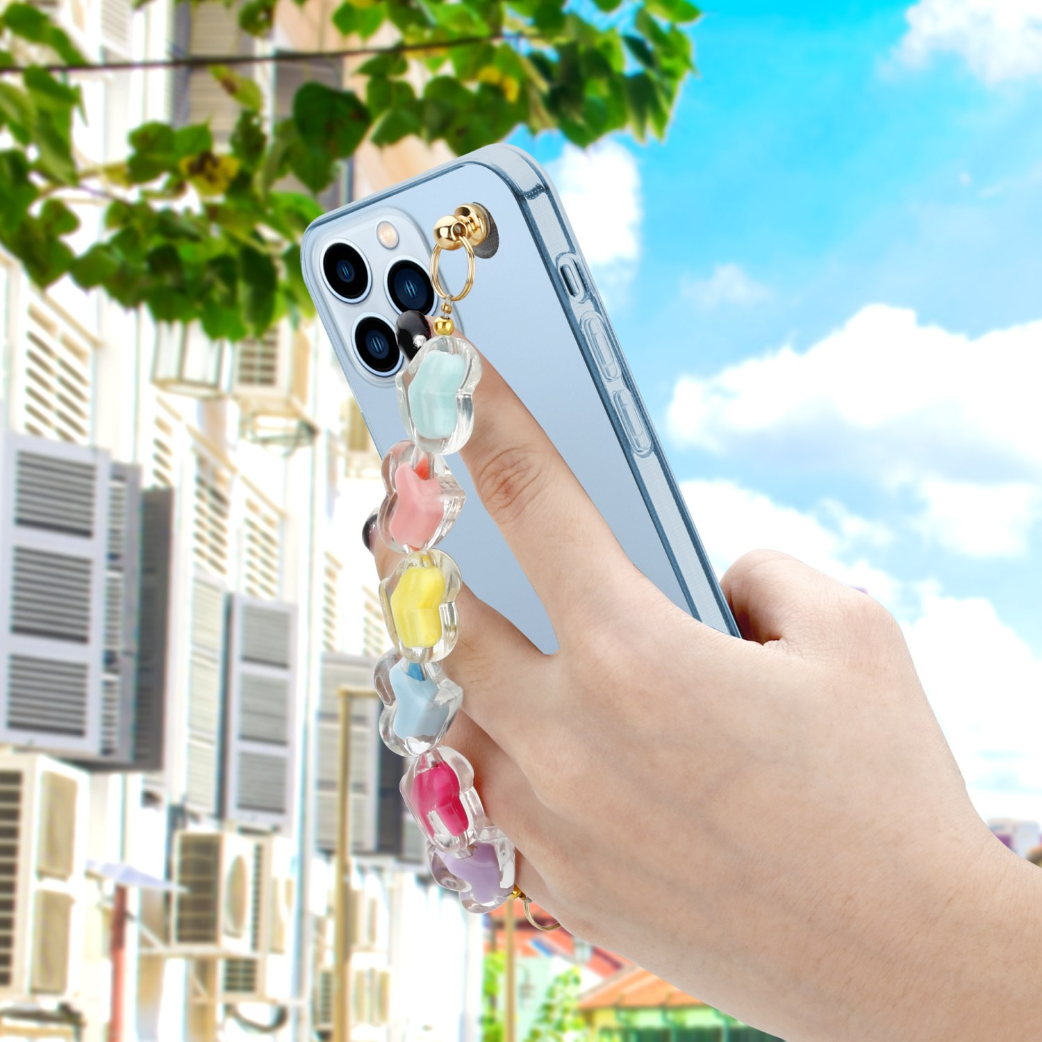 Hand hält hellblaues Telefon mit Blumenanhänger. Hintergrund Gebäude und blauer Himmel.