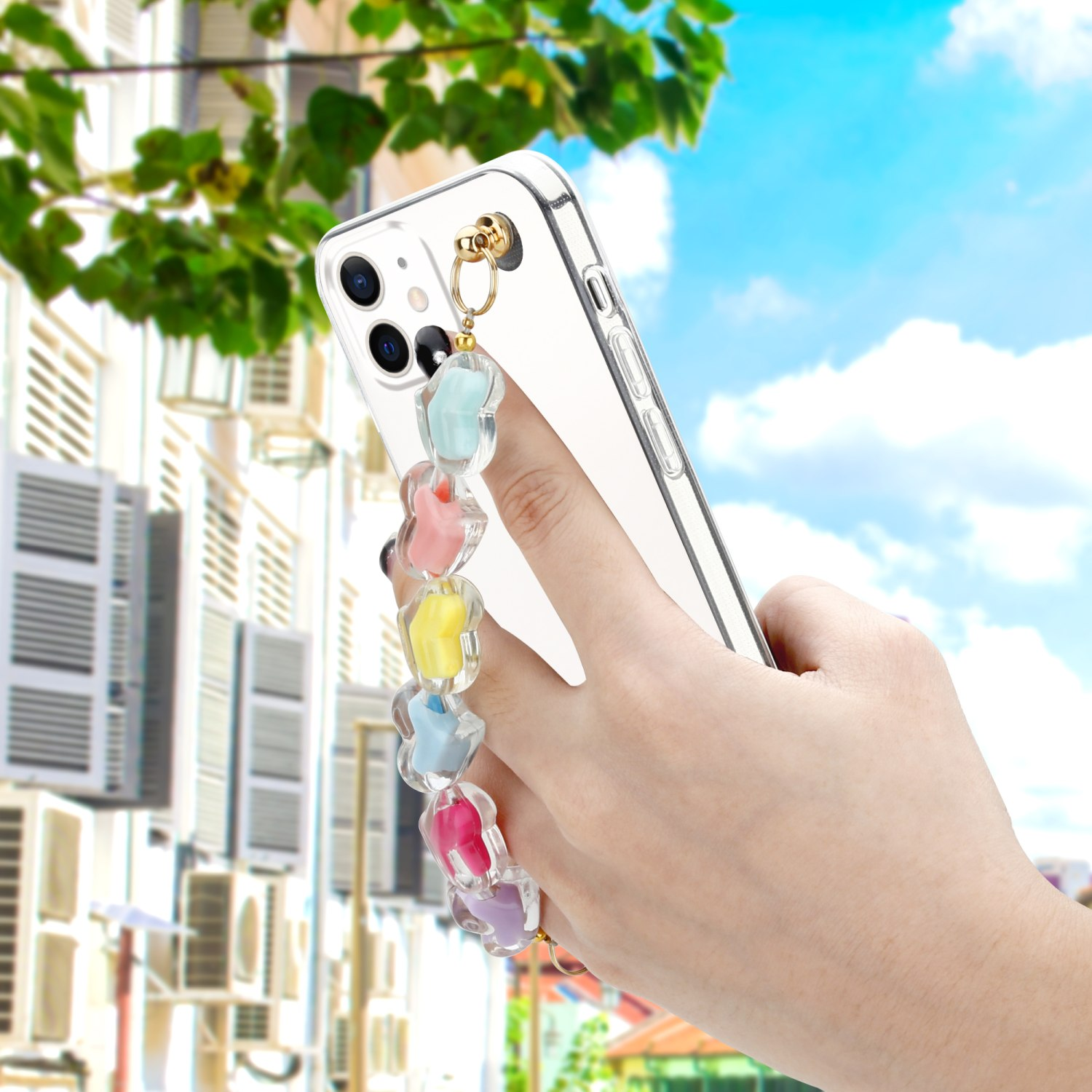 Eine Hand hält ein Telefon mit transparenter Hülle und herzförmiger Kette. Gebäude und blauer Himmel im Hintergrund.